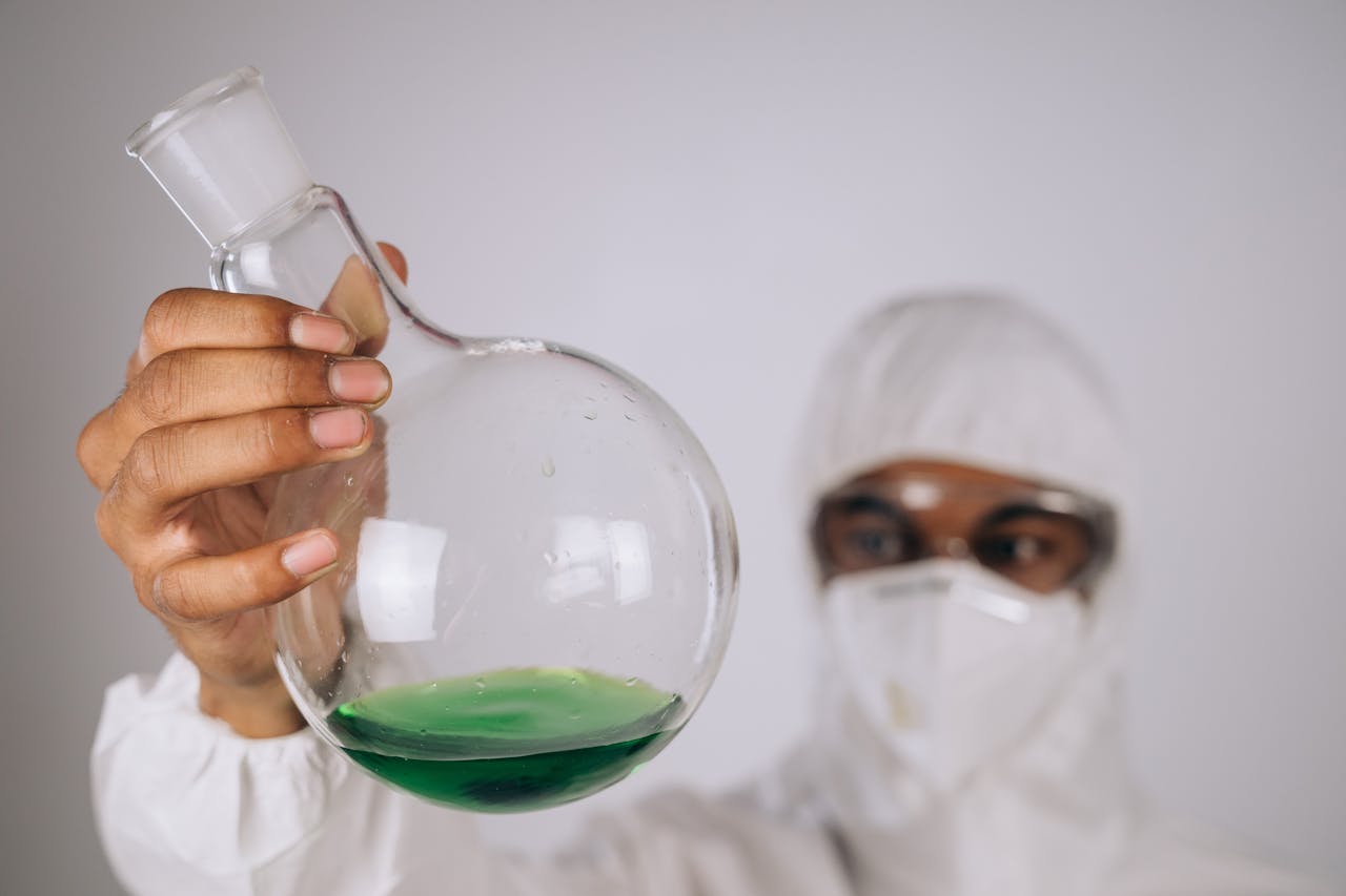 Scientist in protective gear examines green liquid in a flask during a lab experiment.